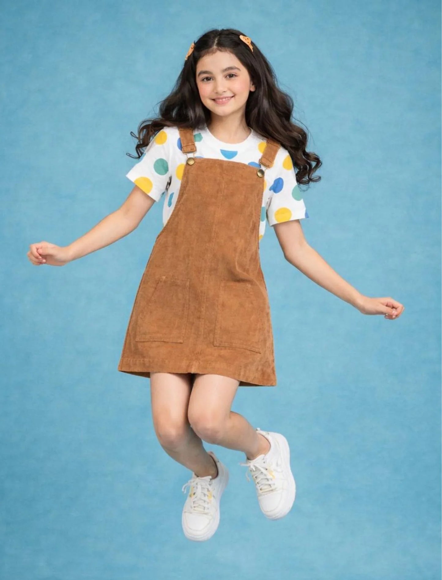 Smiling girl in brown dungaree dress, polka dot t-shirt and white sneakers jumping on blue background