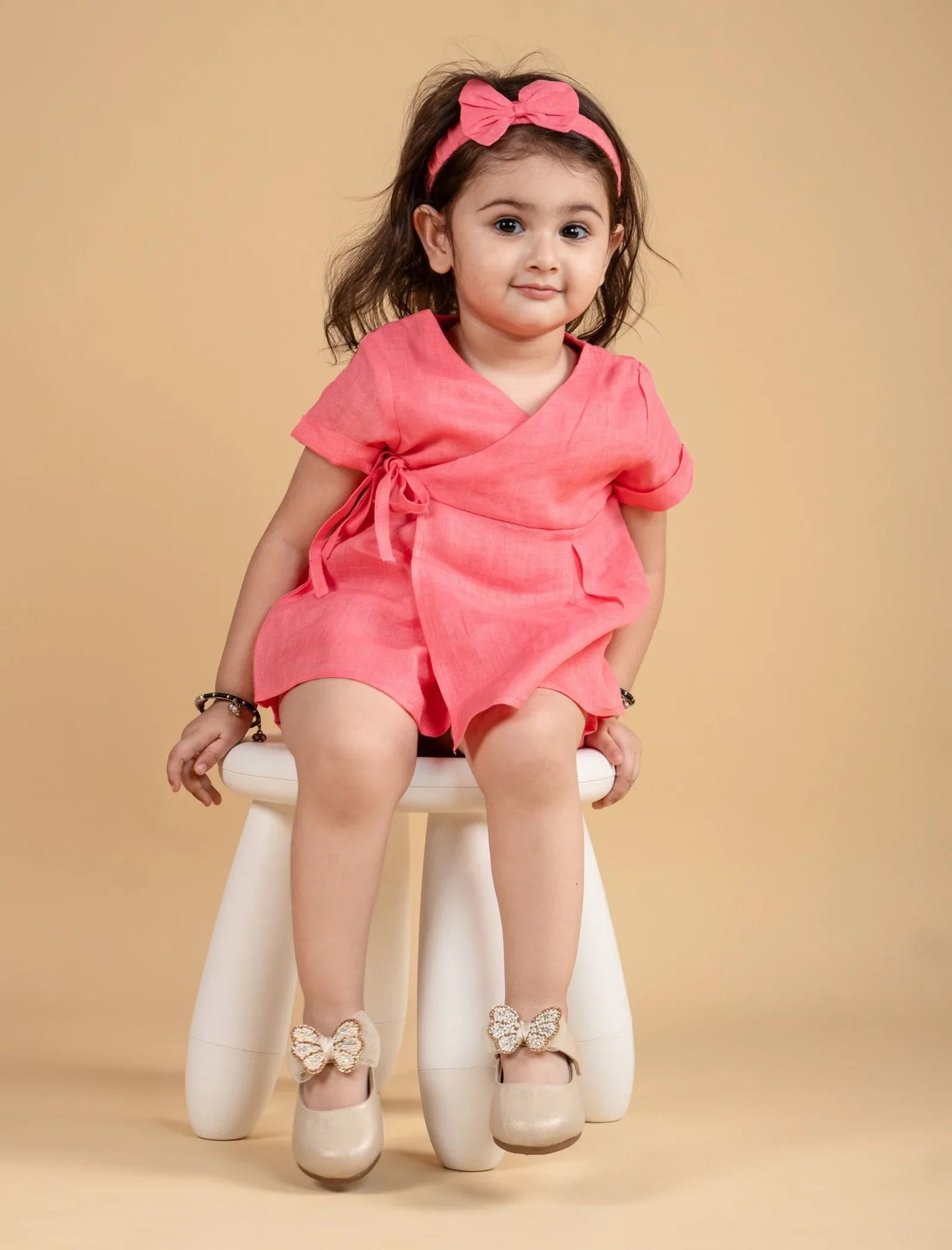 Toddler girl in a pink dress with matching headband, sitting on a stool, cream shoes with bows