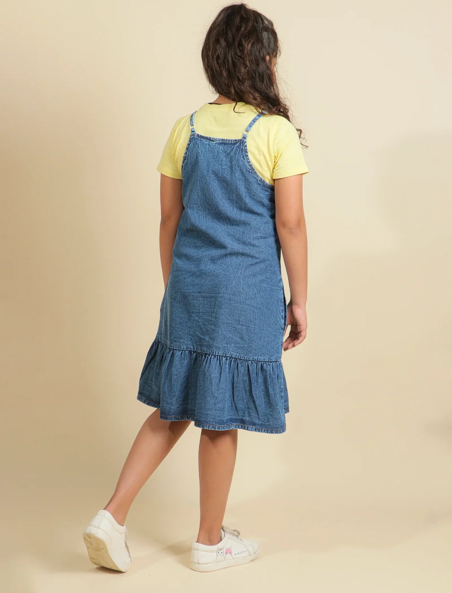 Girls wearing a blue denim pinafore dress over a yellow shirt on a beige background