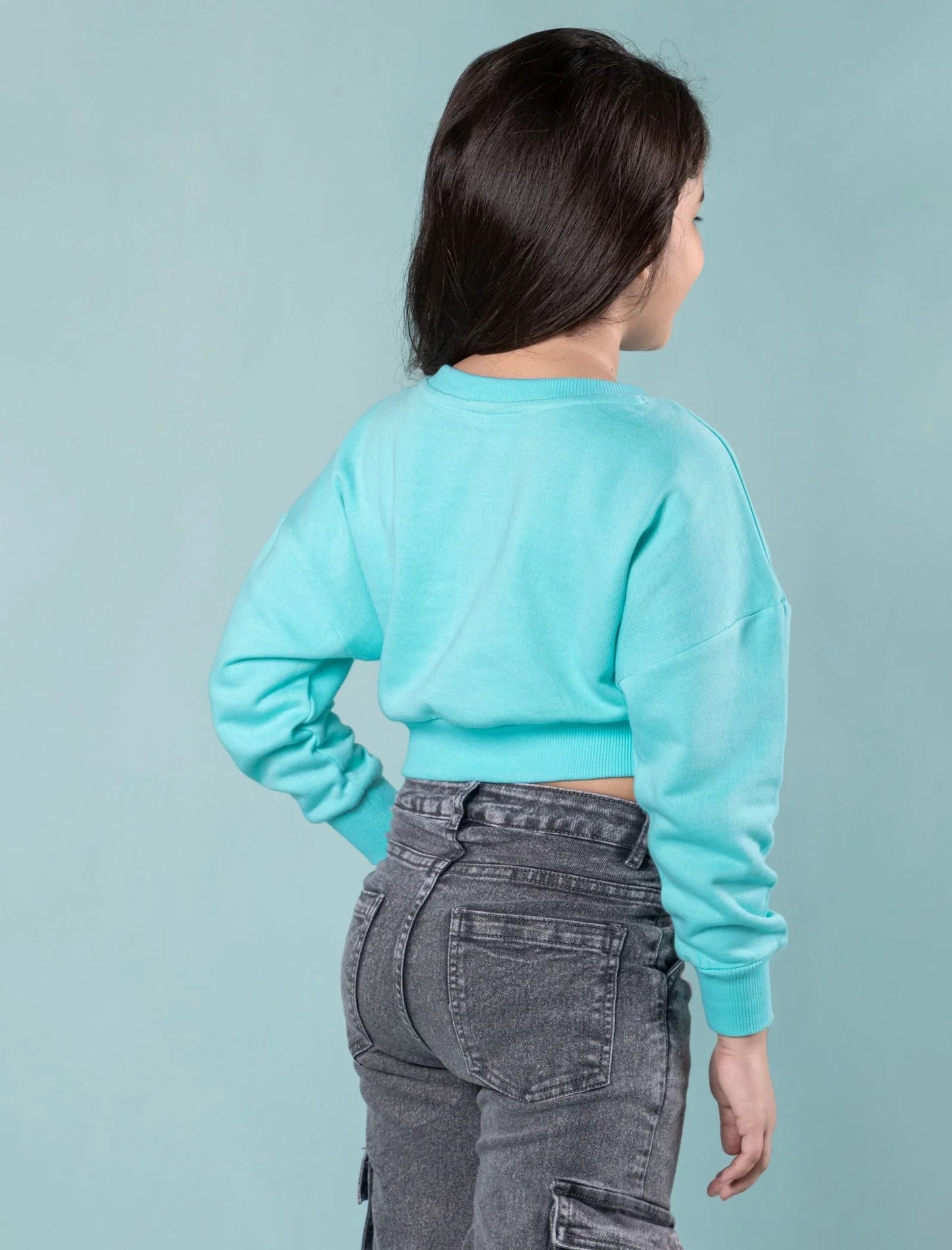 Girl wearing turquoise winter sweatshirt and gray jeans, back view, blue background