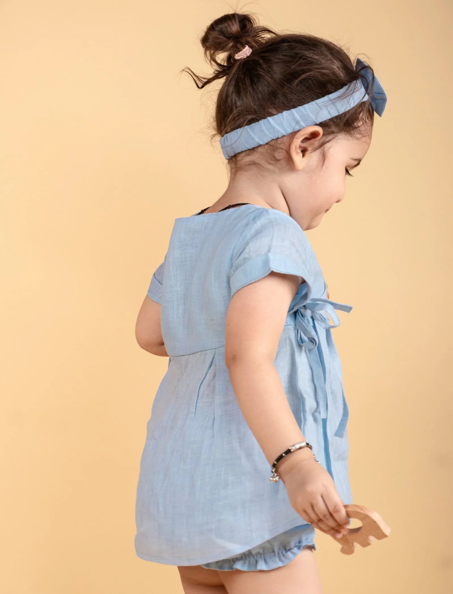 Baby girl in light blue wrap style dress and matching headband, holding a wooden toy.
