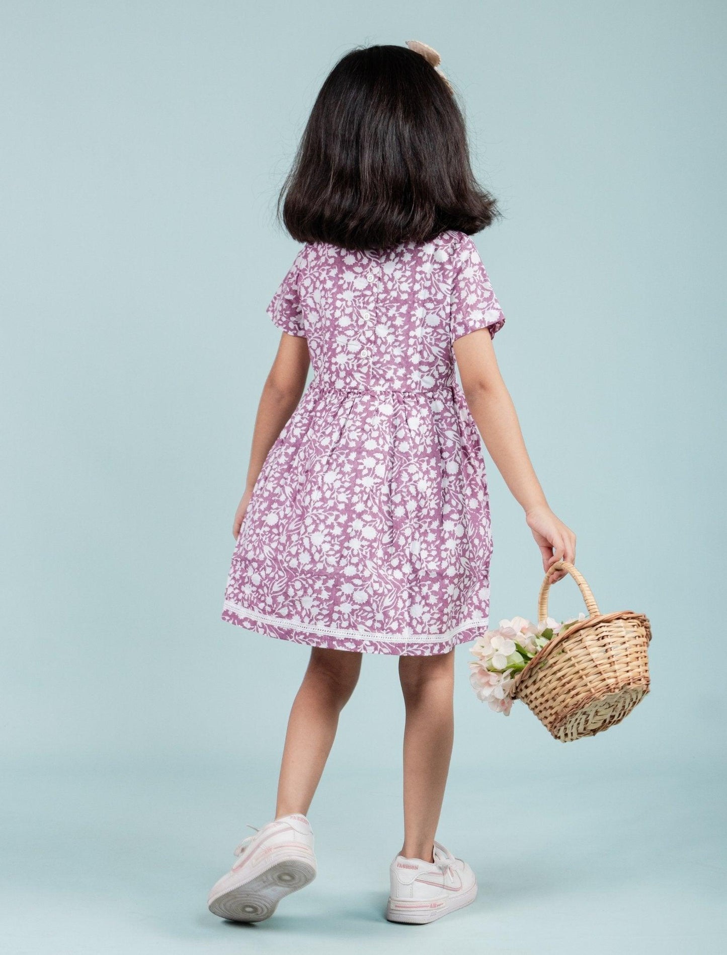 Girl in a pink block printed frock, holding a wicker basket with flowers, pastel background.
