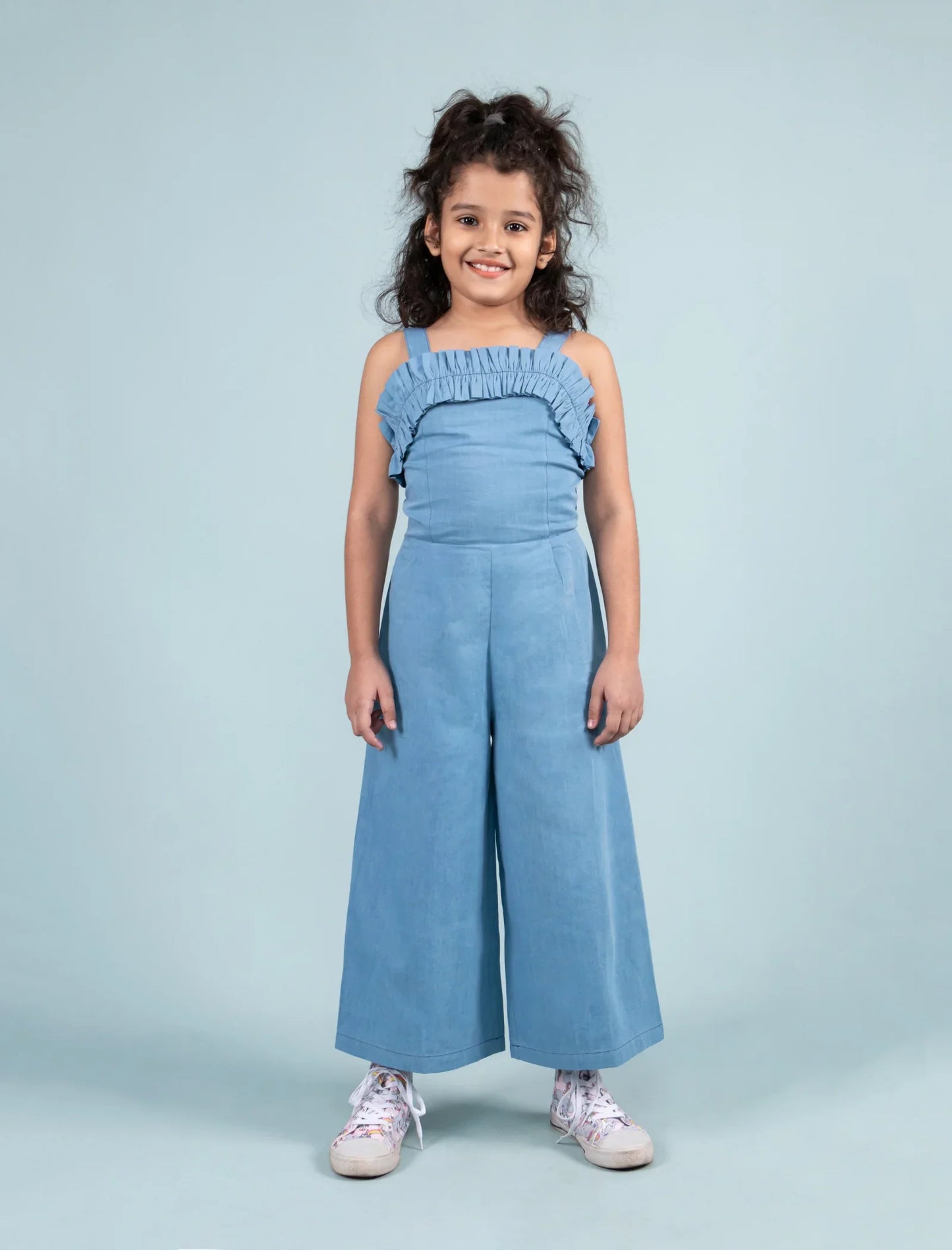 Girl in light blue frilled denim jumpsuit with wide legs and white sneakers, studio background