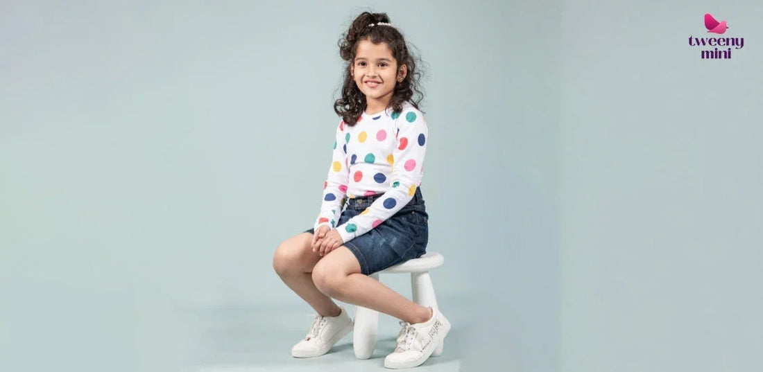 Smiling girl in polka dot top and denim skirt, sitting on a stool, kids wear fashion