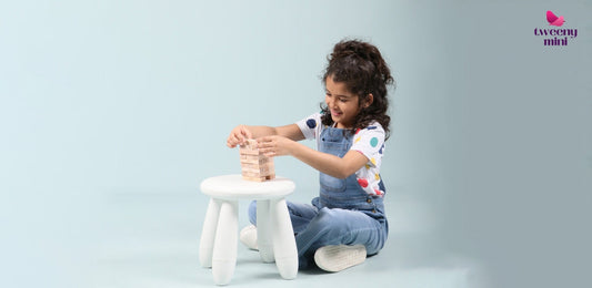 Smiling girl in denim overalls and polka dot t-shirt playing Jenga on white stool