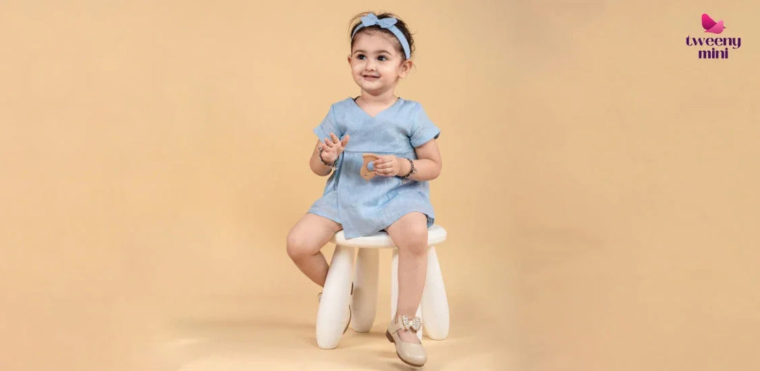 Smiling young girl in blue short-sleeve dress and headband sitting on white stool against beige background