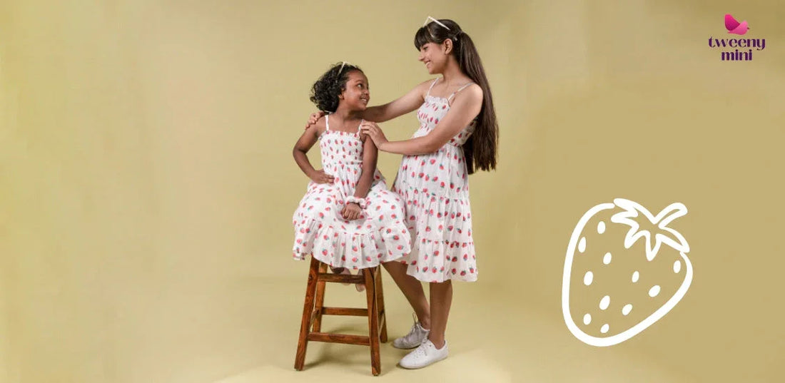 Girls in summer dresses with strawberry print, one seated on a stool, smiling on beige background