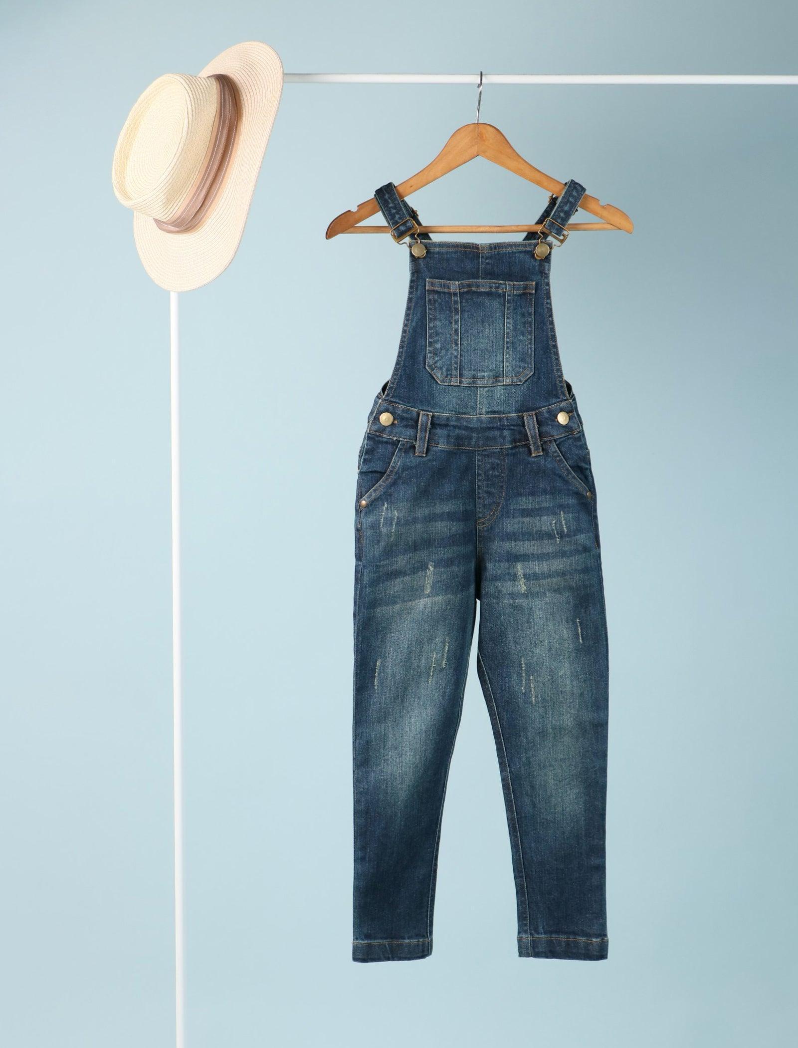 Kids denim dungaree with bib pocket on hanger and straw hat against light blue background