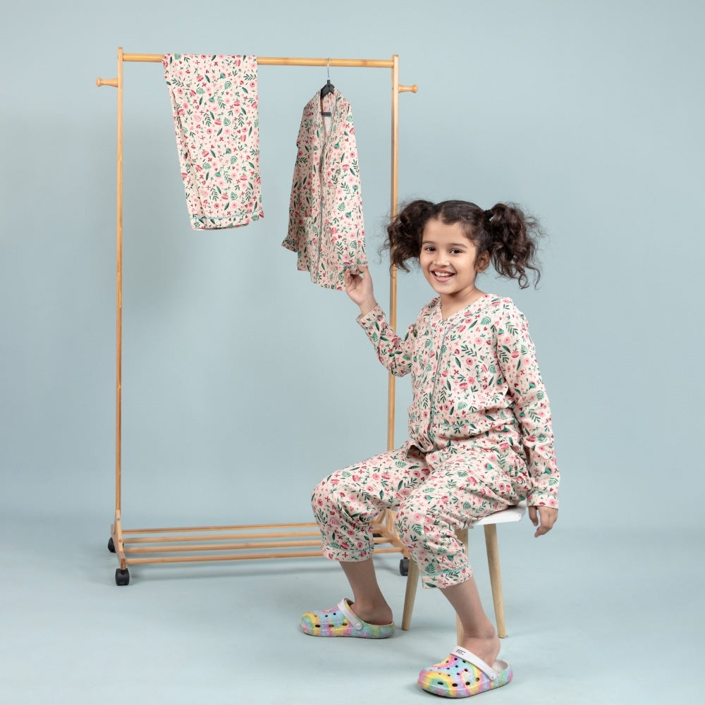 Smiling girl in floral print night suit sitting on stool, kids sleepwear set displayed on rack.