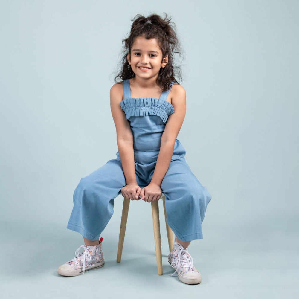 Smiling girl in blue sleeveless jumpsuit with ruffle detail, sitting on a stool, kids fashion