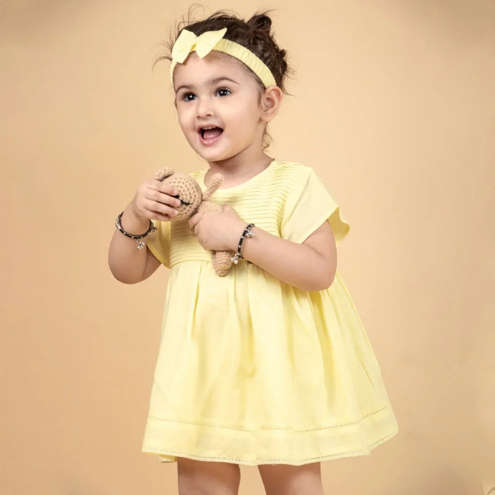 Smiling baby girl in yellow dress and headband holding a stuffed toy against beige background.