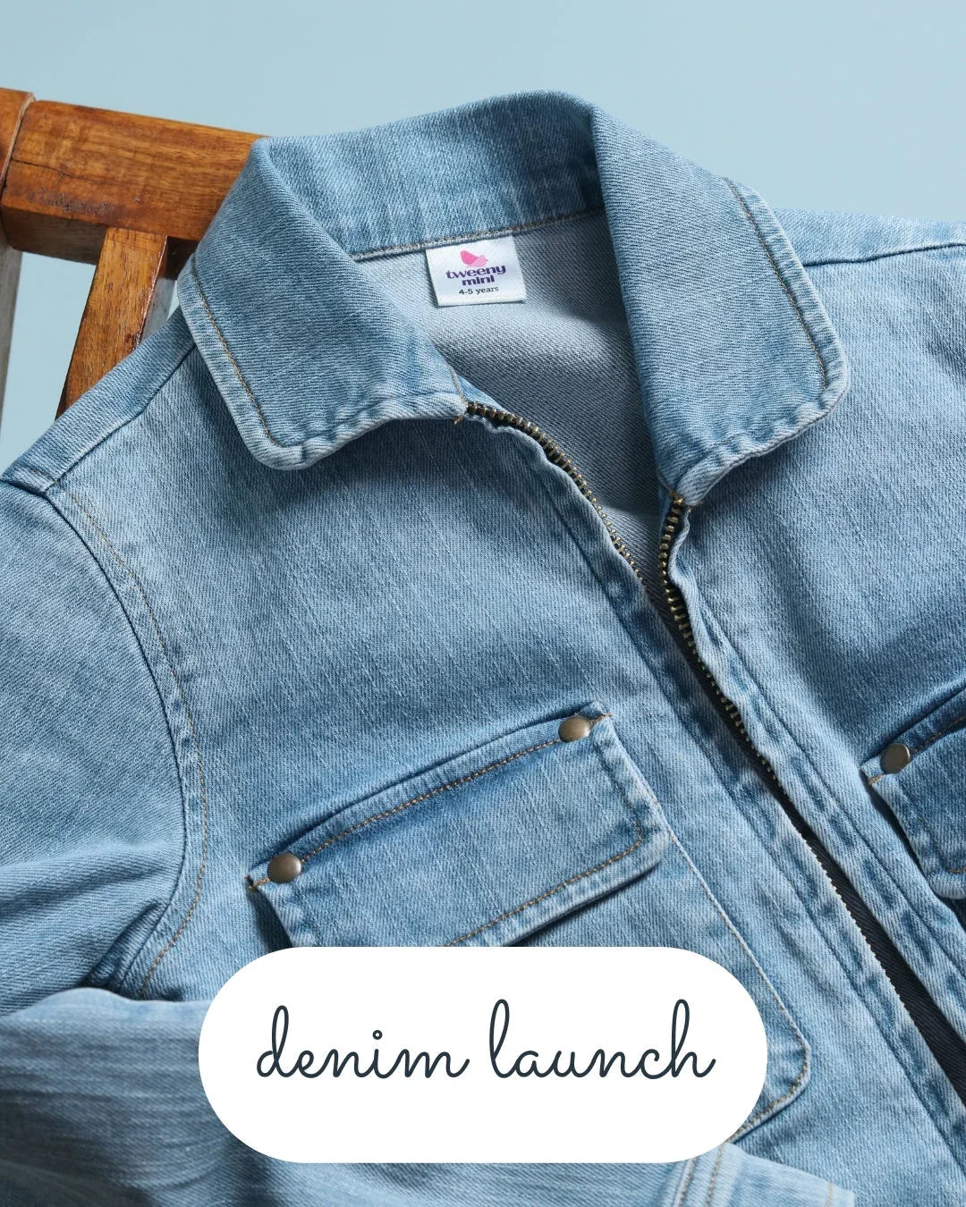 Light blue kids denim jacket with front pockets and zipper, displayed on a wooden chair.