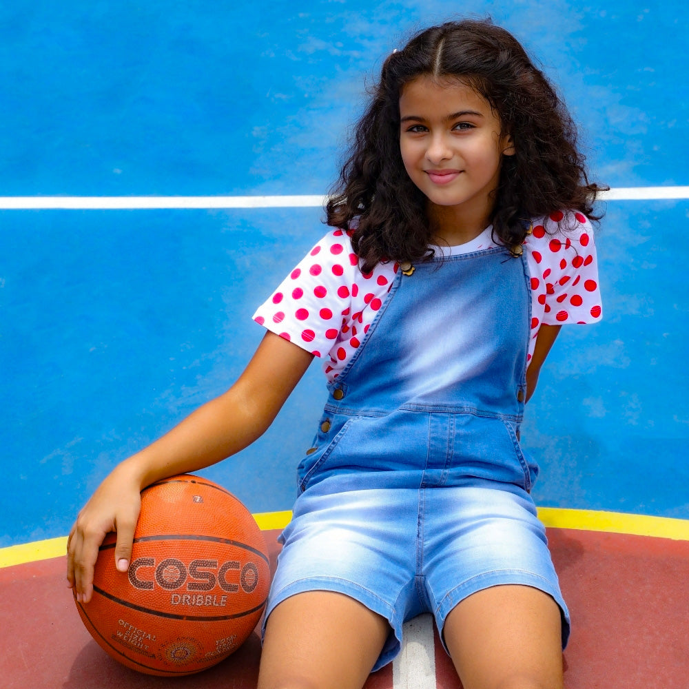 Girl in denim dungaree shorts and polka dot t-shirt sitting on a basketball court with a basketball
