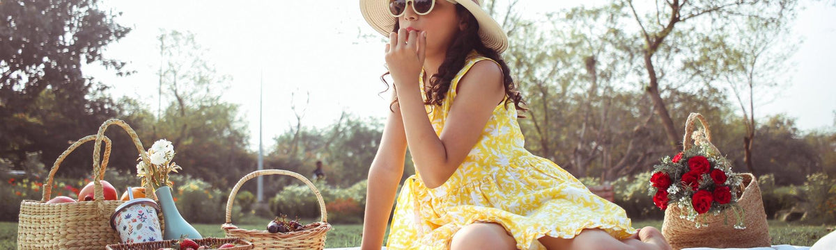 Girl in yellow floral dress and sun hat having summer picnic on grass with baskets and flowers.