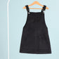 Black pinafore dress on a hanger against a light blue background