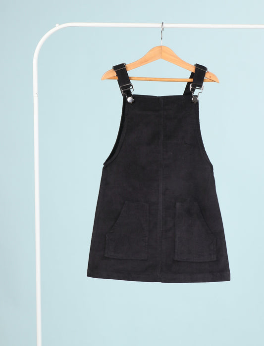 Black pinafore dress on a hanger against a light blue background