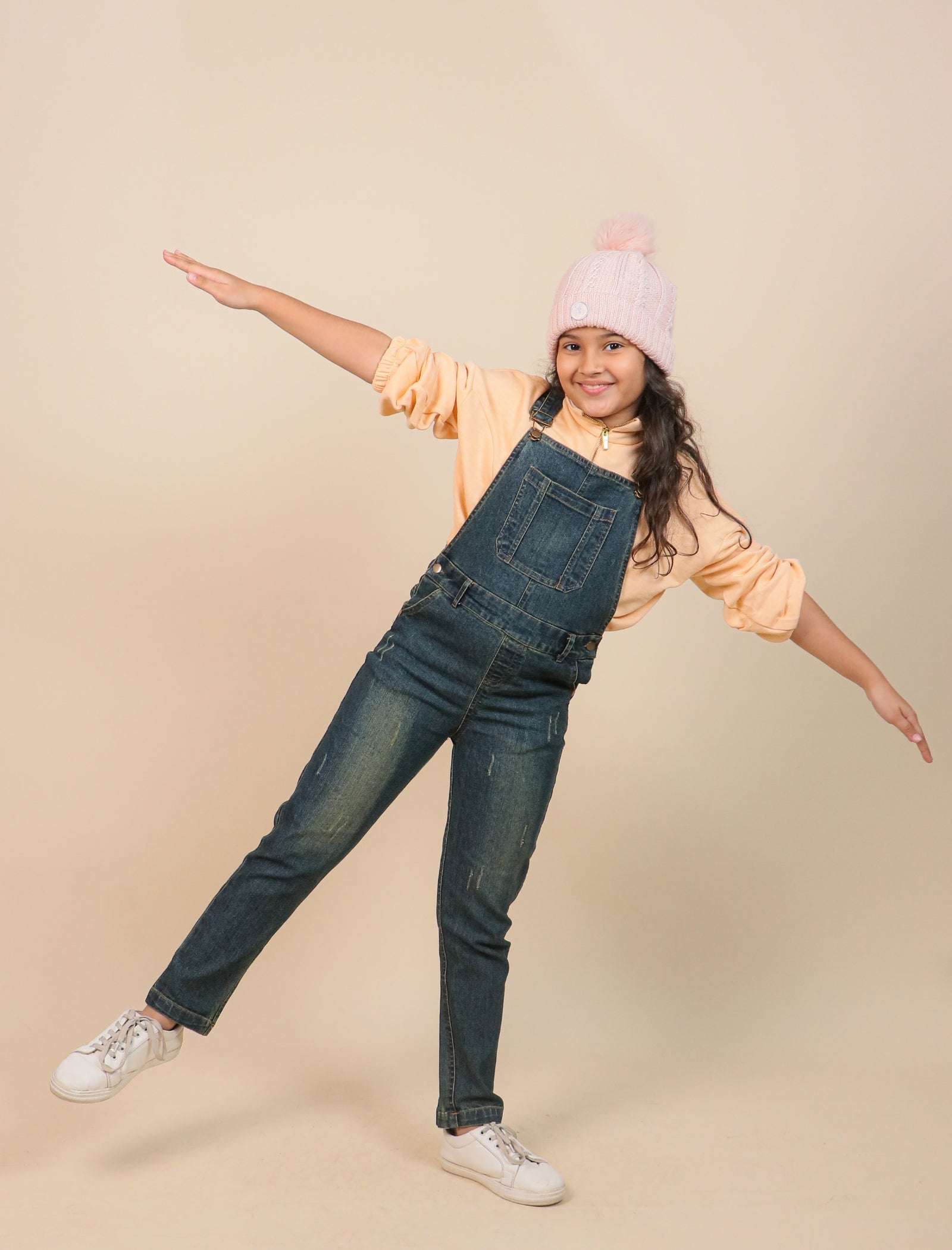 Girls wearing denim overalls and a light pink beanie on a beige background