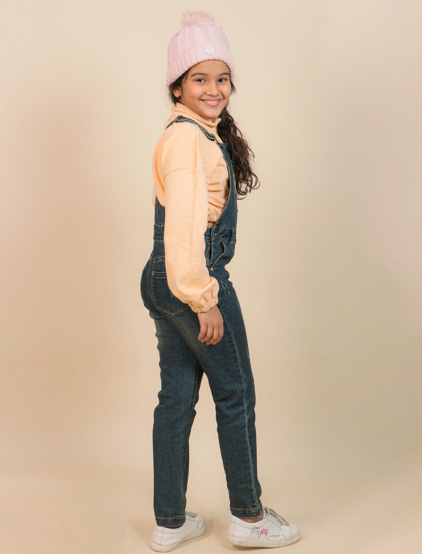 Girls wearing a pink beanie, light orange shirt, and blue denim overalls on a beige background