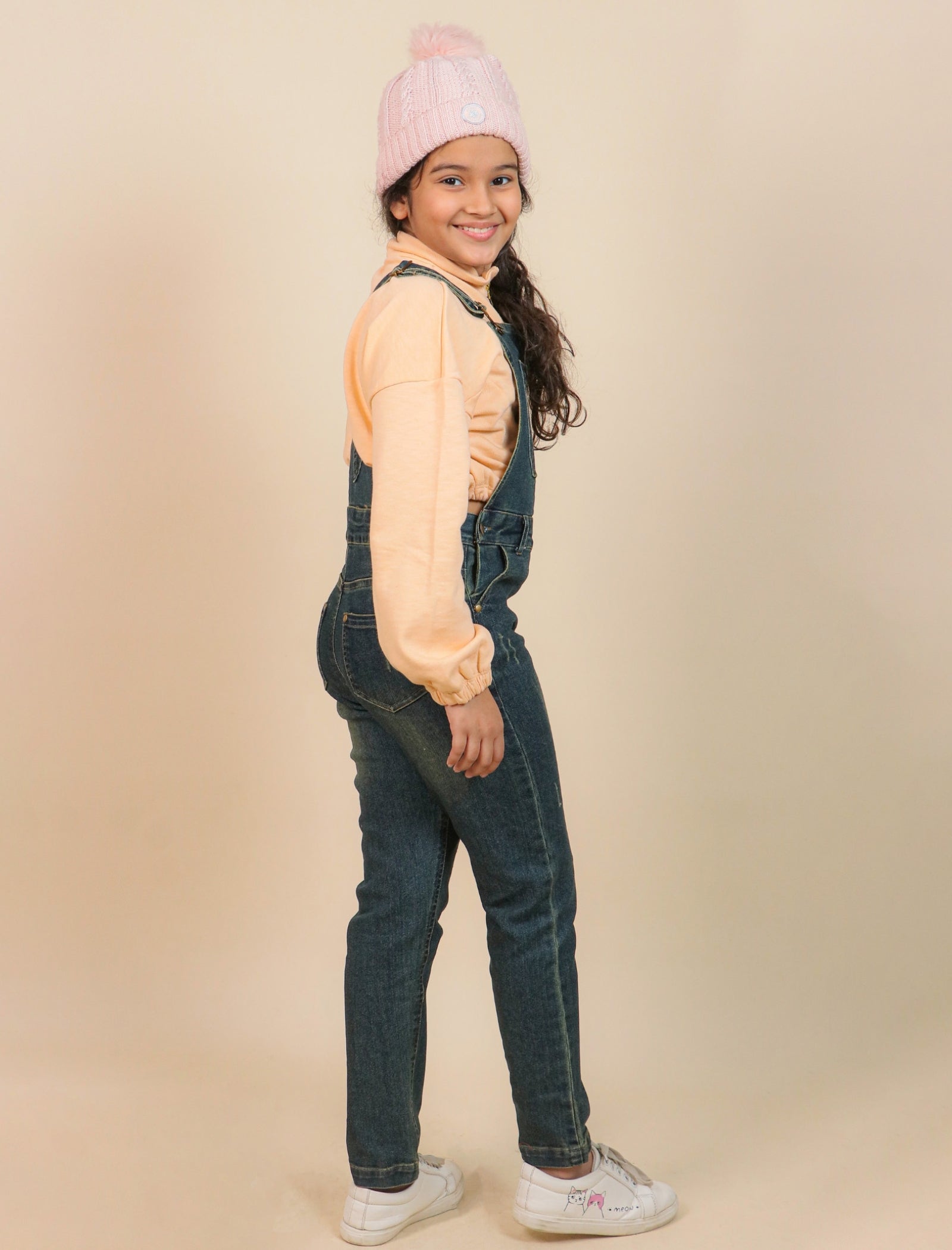 Girls wearing a pink beanie, light orange shirt, and blue denim overalls on a beige background