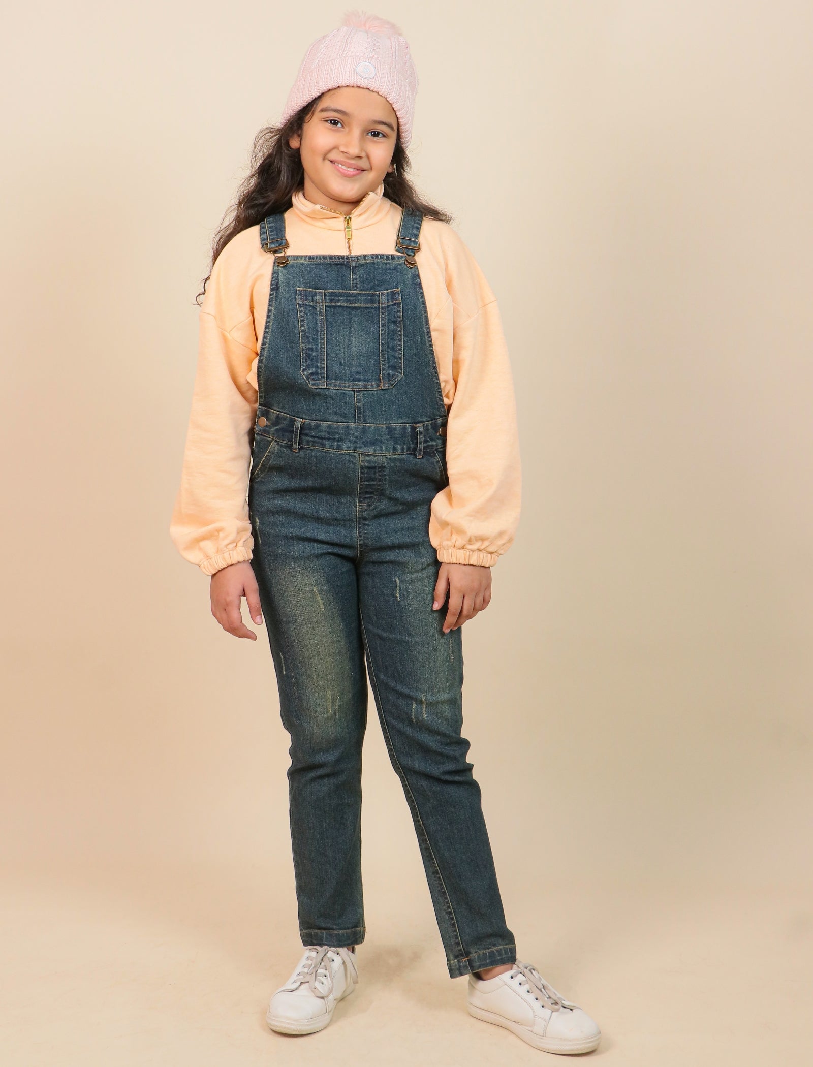Girls wearing a peach jacket, denim overalls, and white sneakers on a beige background