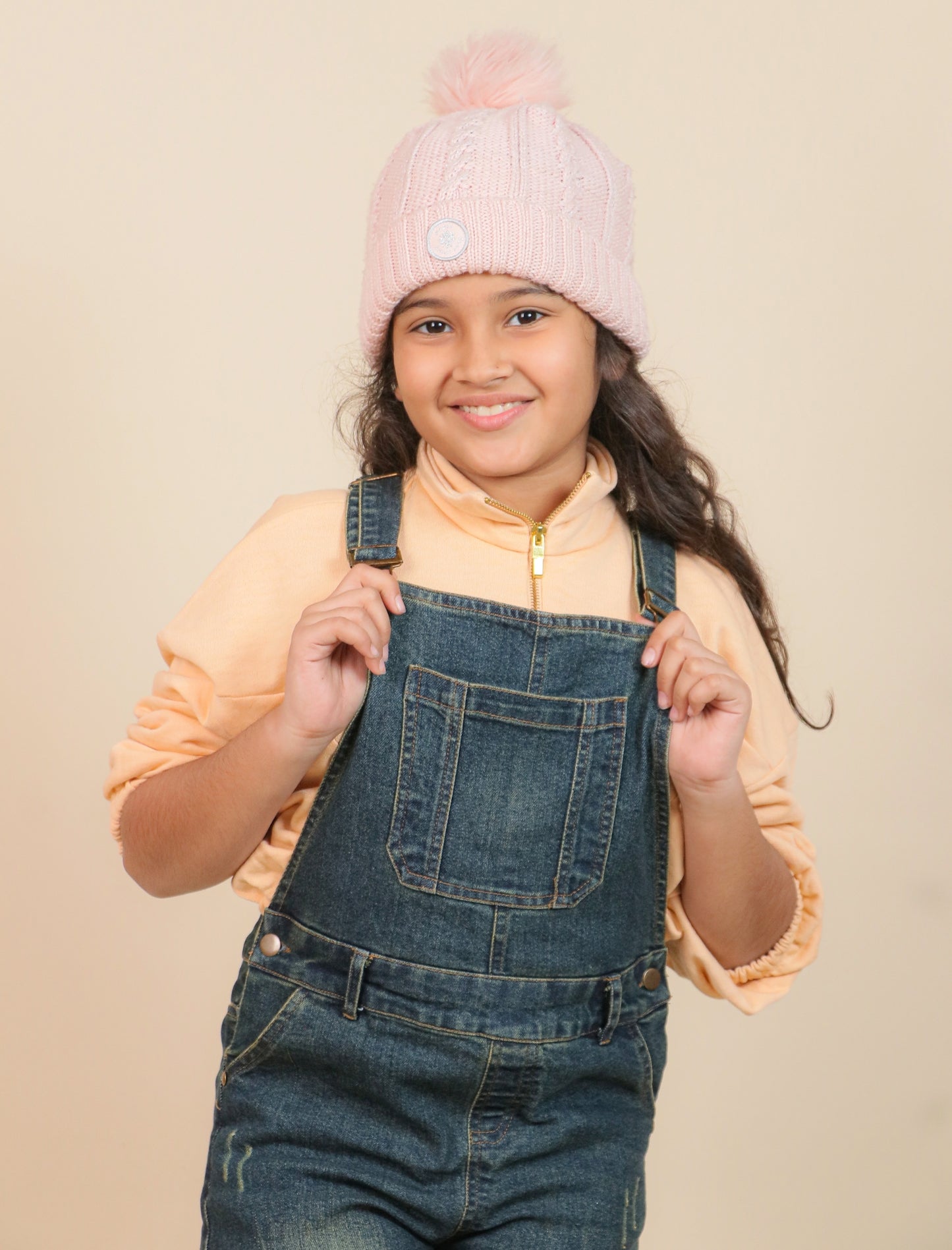 girls wearing a pink knit hat and denim overalls against a beige background