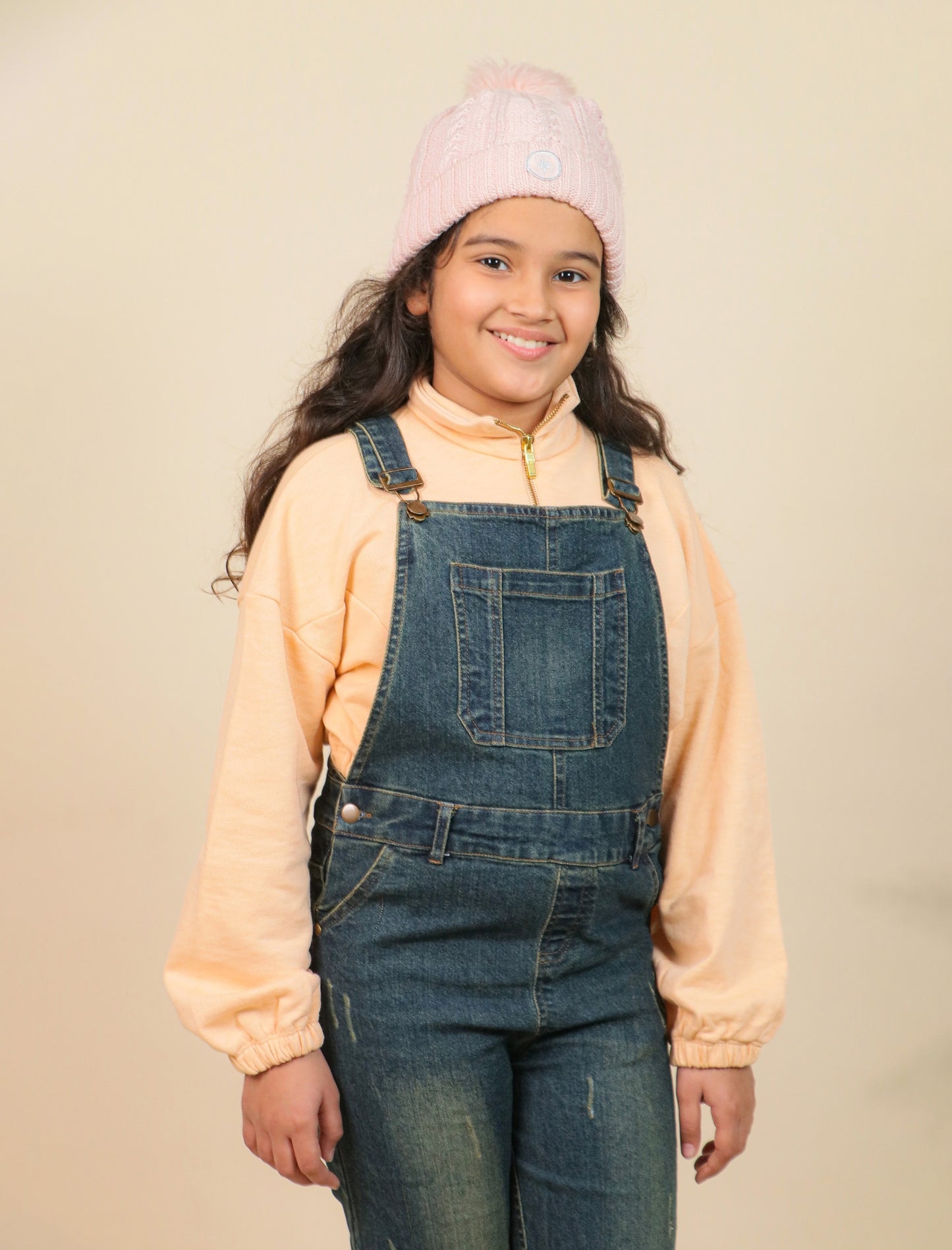 Girls wearing a pink beanie and denim overalls against a beige background
