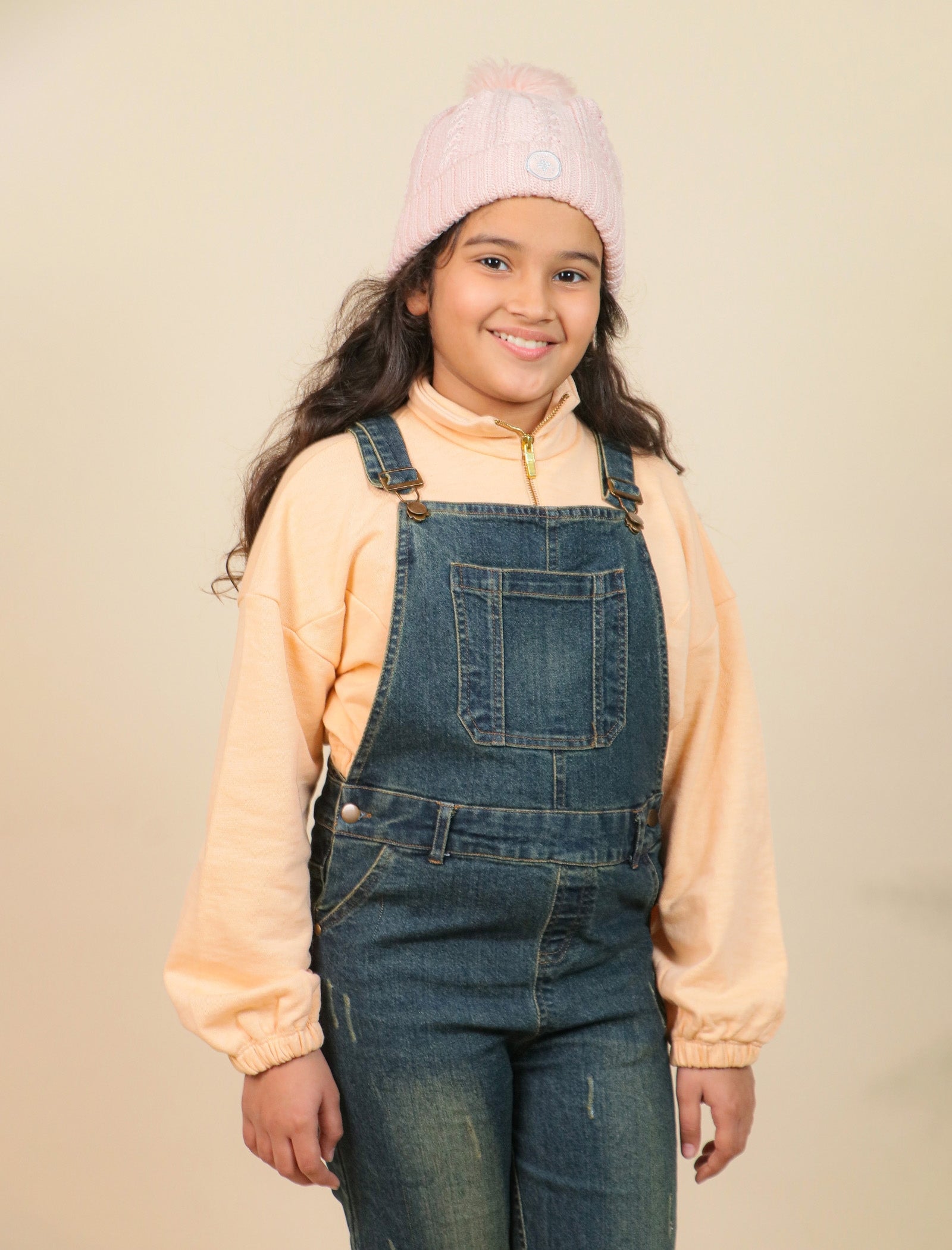 Girls wearing a pink beanie and denim overalls against a beige background