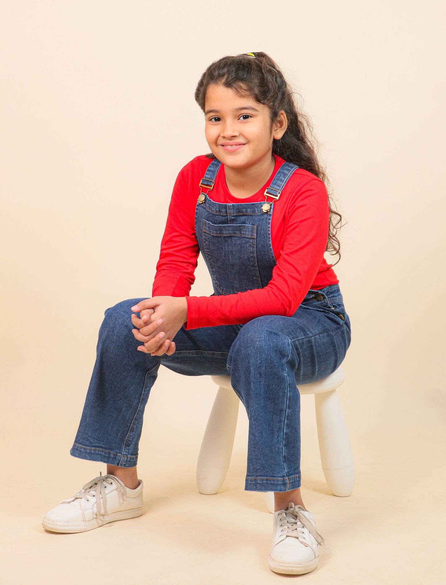 Young girl wearing a red long-sleeve shirt and blue denim overalls sitting on a white stool against a beige background.