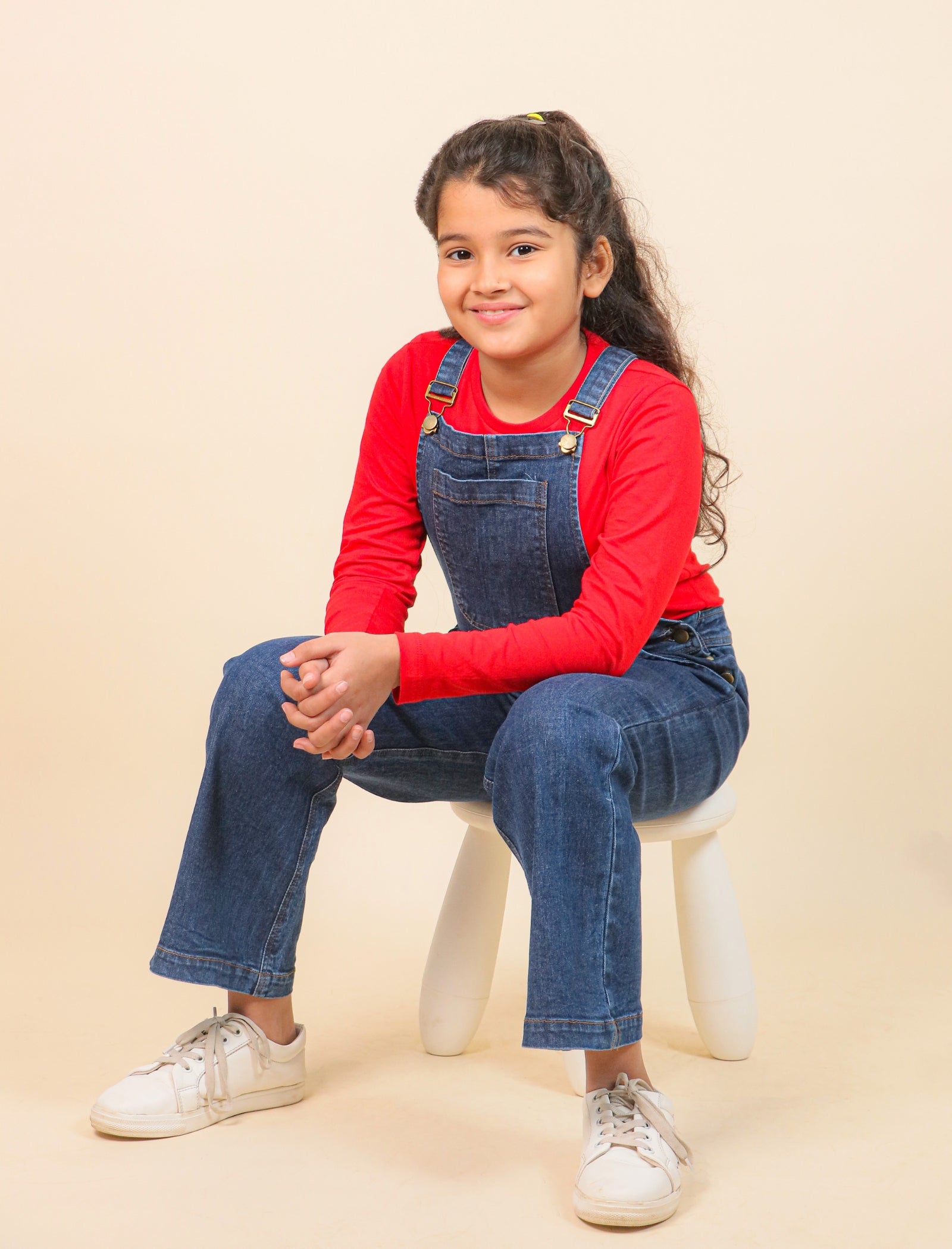 Young girl wearing a red long-sleeve shirt and blue denim overalls sitting on a white stool against a beige background.