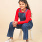 Girls wearing a red long-sleeve shirt and blue denim overalls sitting on a white stool against a beige background.