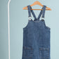 Blue denim pinafore dress on a hanger against a light blue background