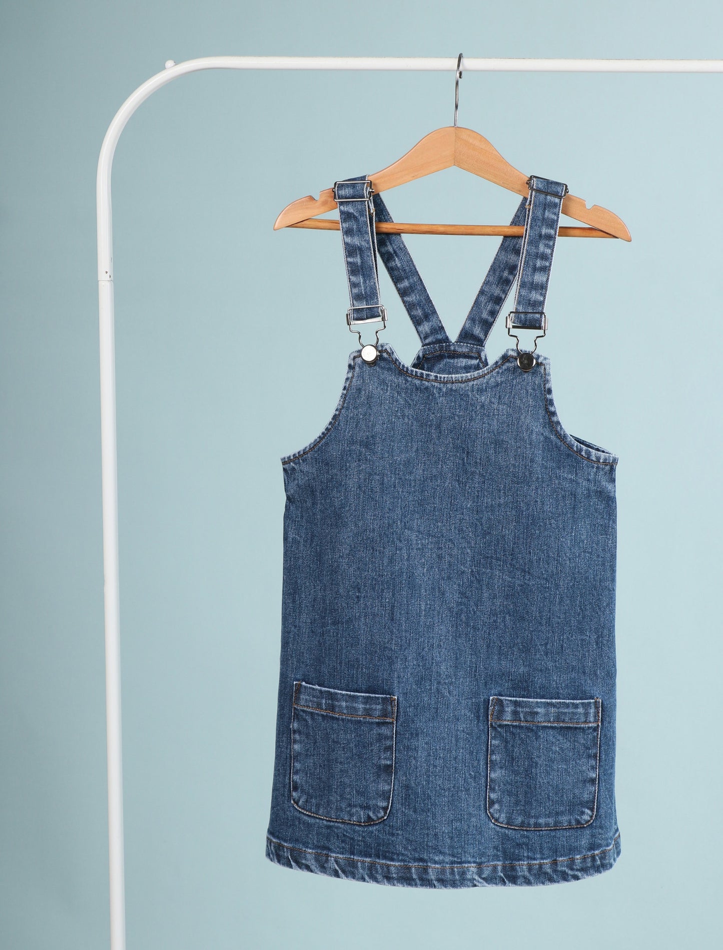 Blue denim pinafore dress on a hanger against a light blue background