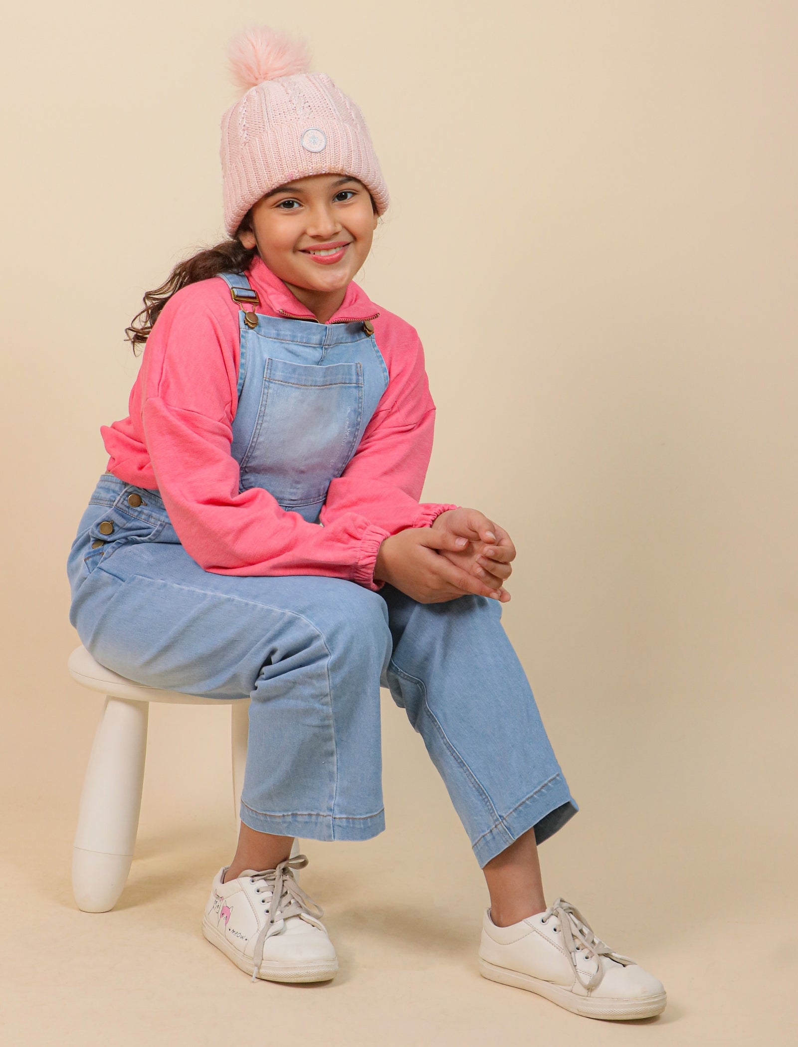 Girls wearing a pink hoodie, denim overalls, and white sneakers on a beige background
