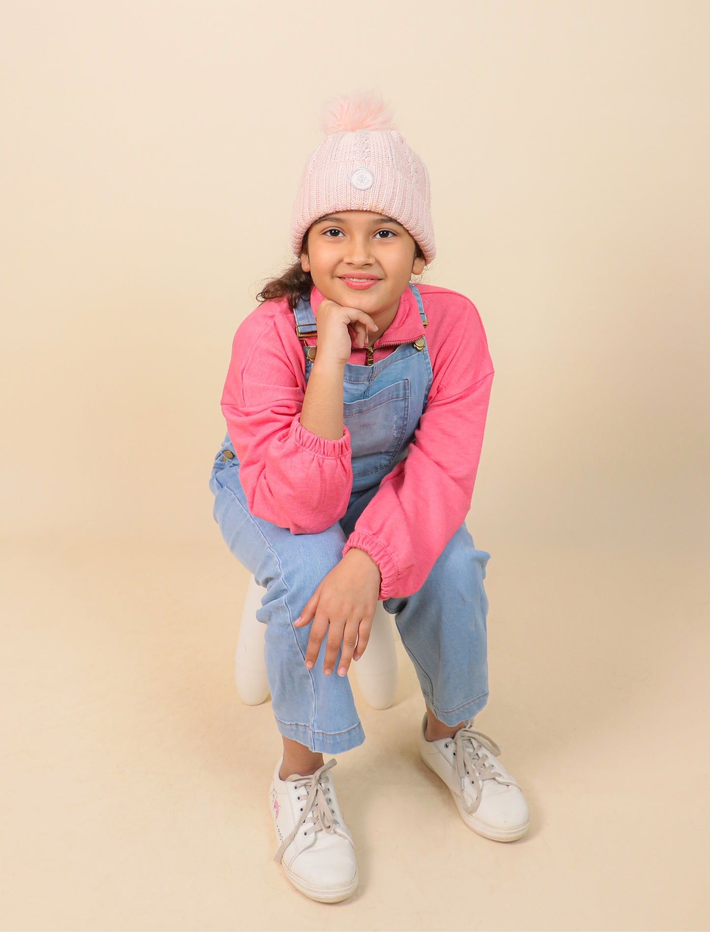 Girls wearing a pink jacket, denim overalls, and white sneakers on a beige background