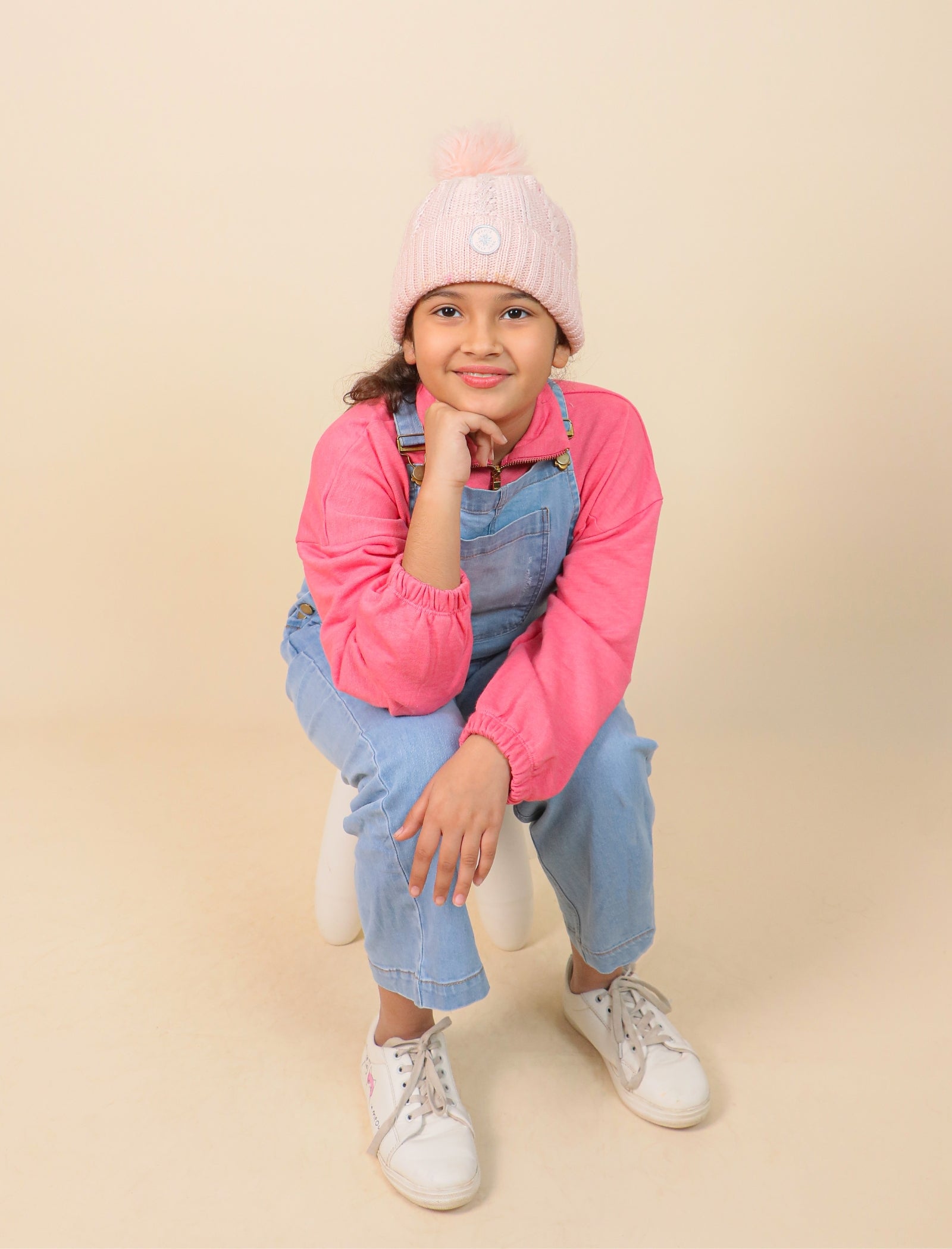 Girls wearing a pink jacket, denim overalls, and white sneakers on a beige background