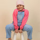 Girls wearing a pink jacket and blue overalls sitting on a white stool against a beige background
