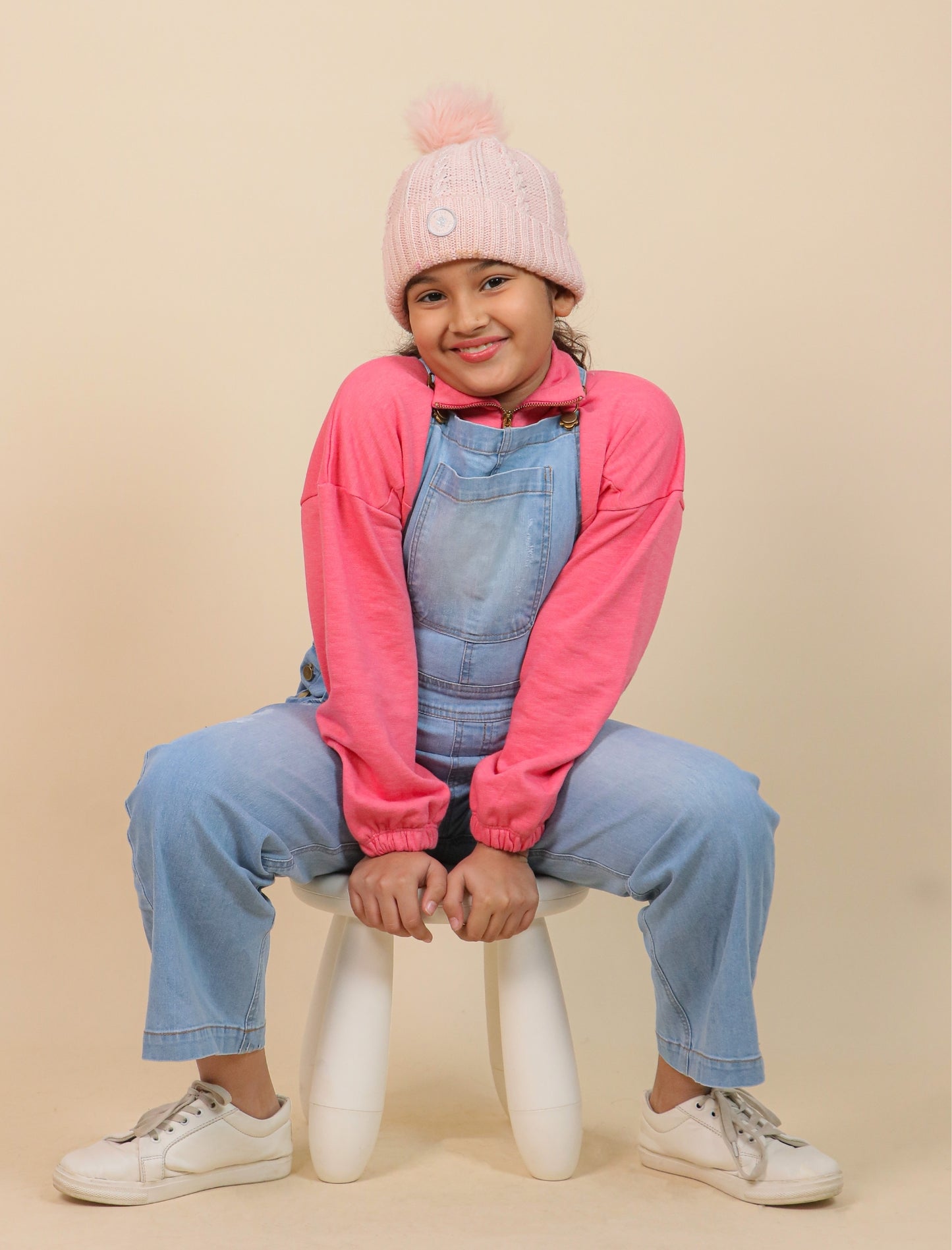 Girls wearing a pink jacket and blue overalls sitting on a white stool against a beige background