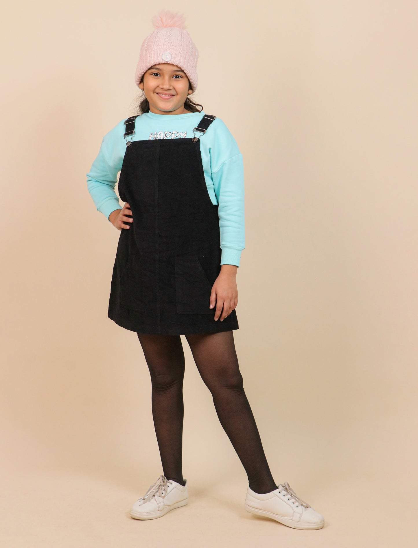 Girls wearing a black pinafore dress, light blue sweatshirt, pink beanie, and white sneakers on a beige background