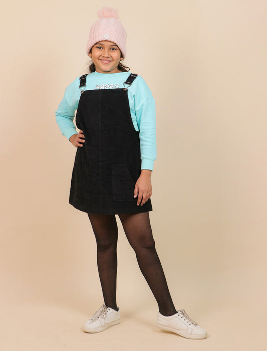 Girls wearing a black pinafore dress, light blue sweatshirt, pink beanie, and white sneakers on a beige background