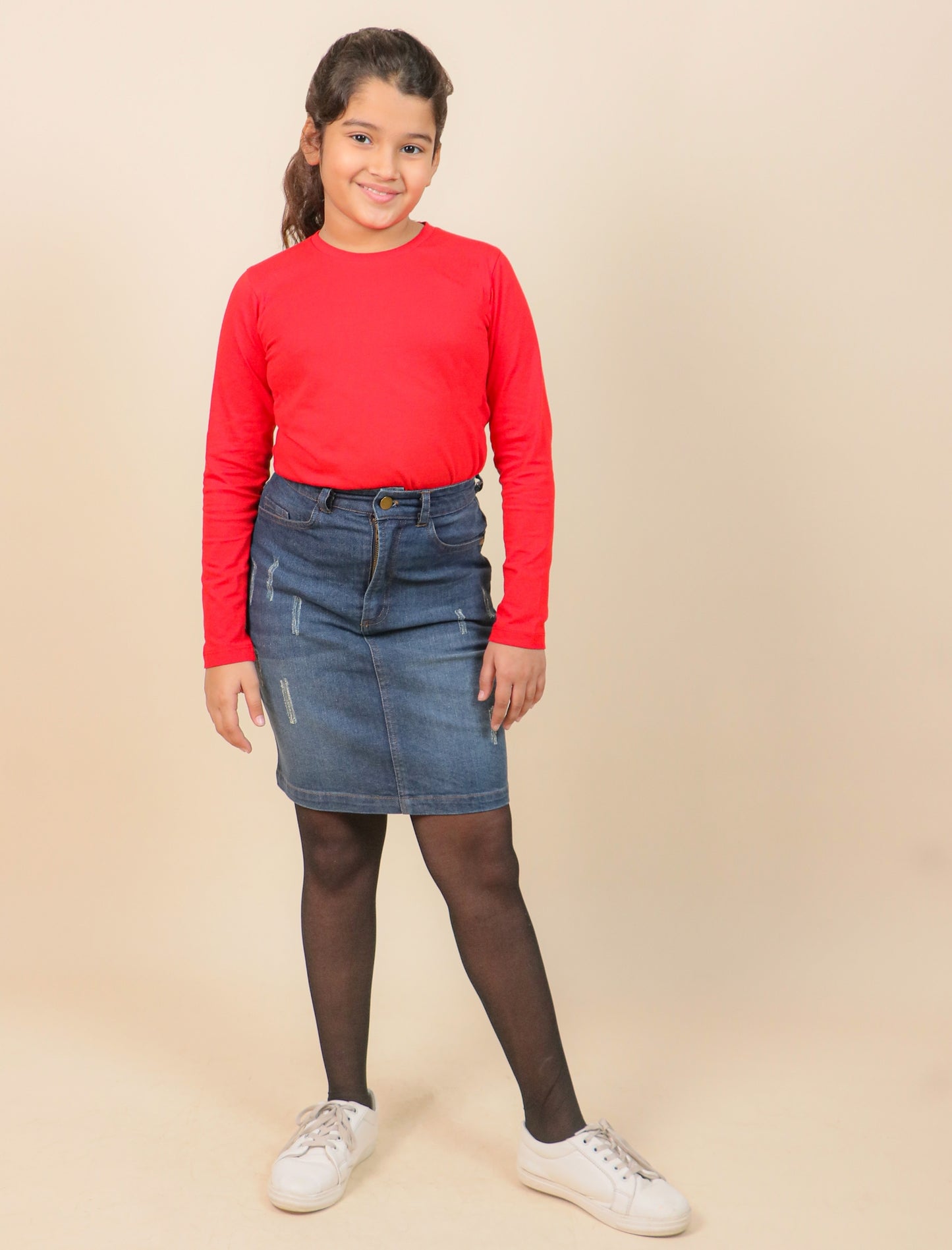 Girl wearing a red sweater, blue denim skirt, and white sneakers on a beige background