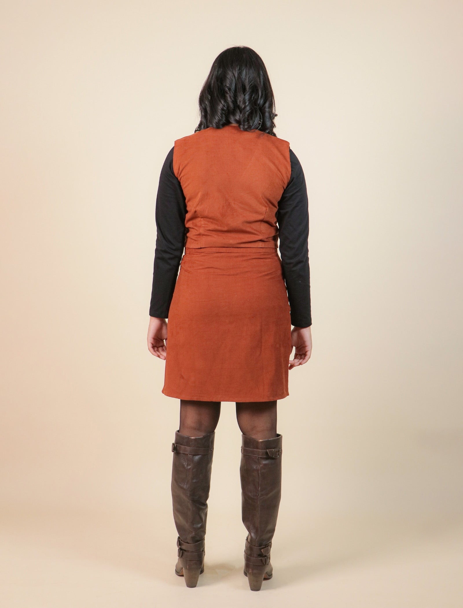 Girls wearing a brown dress with a vest over a black shirt, standing against a beige background
