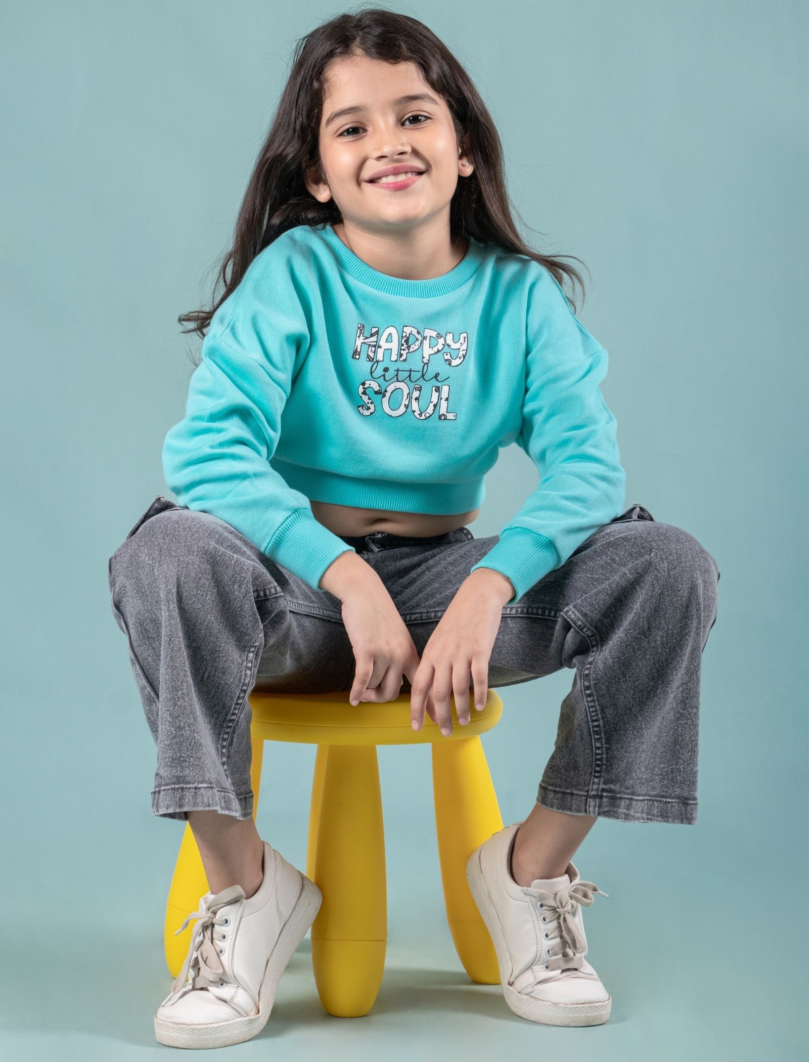 Kids wearing a turquoise sweatshirt with text, sitting on a yellow stool against a light blue background