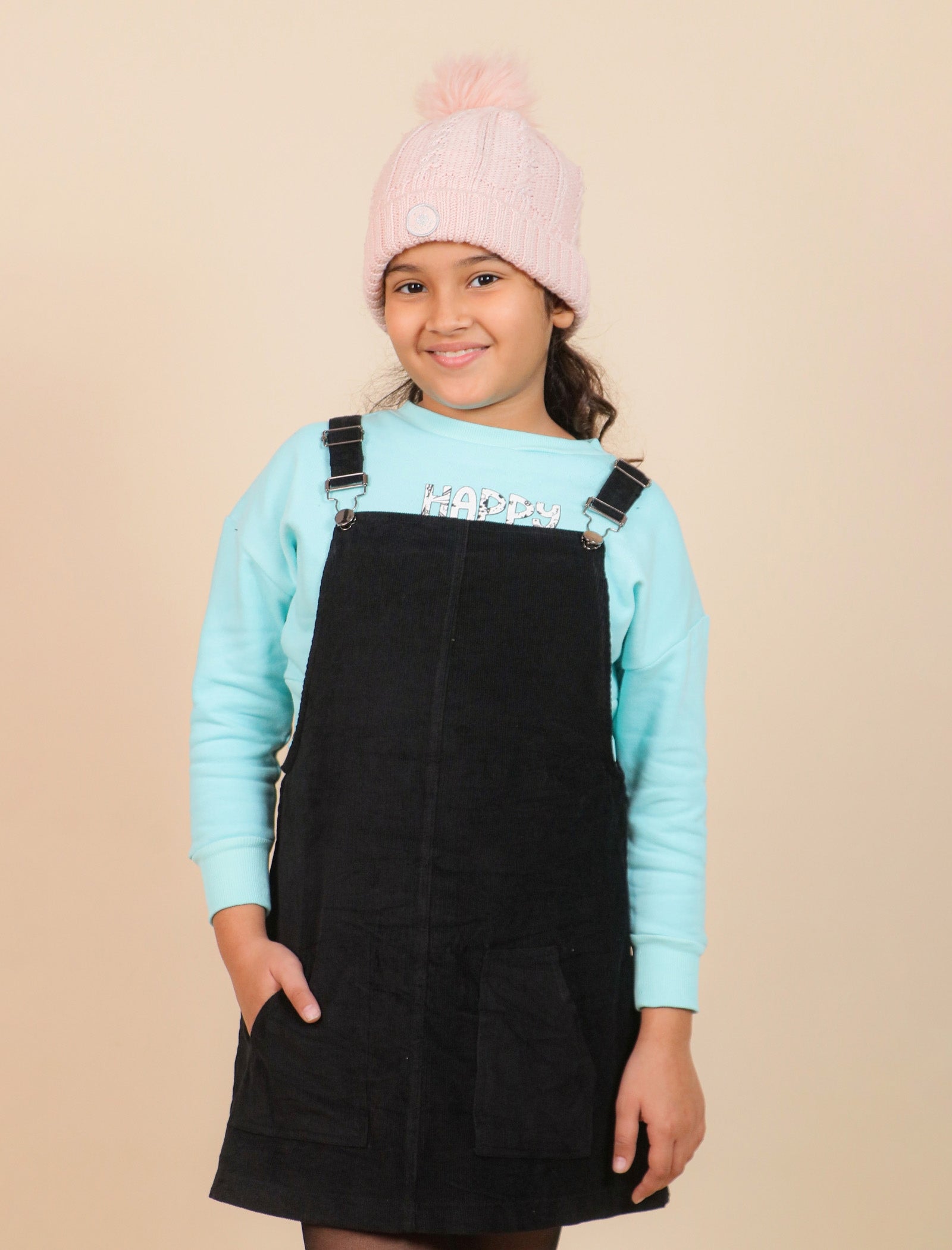 girls wearing a black pinafore dress over a light blue long-sleeve shirt with a pink beanie on a beige background