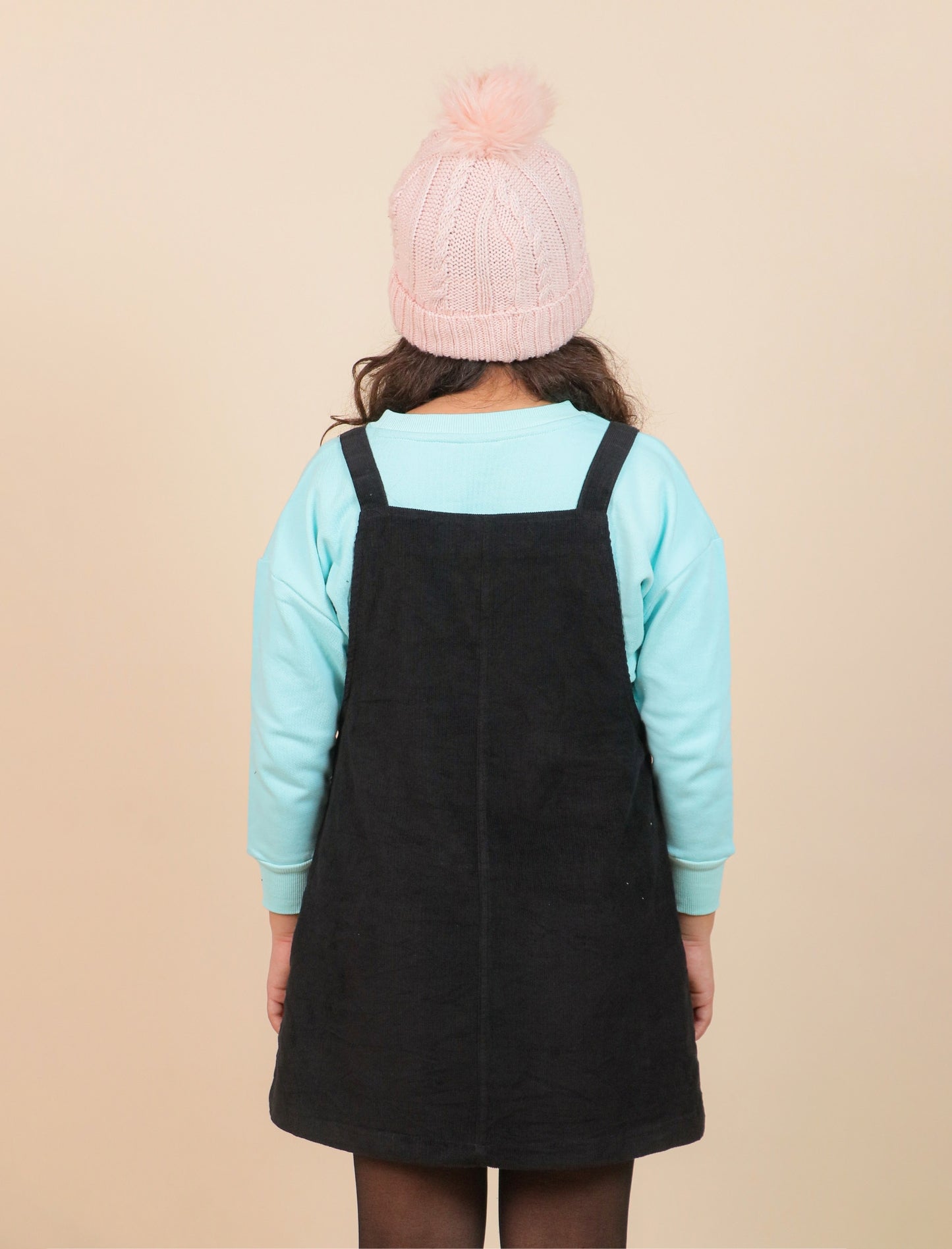 Girls wearing a black pinafore dress over a light blue long-sleeve shirt with a pink knit hat on a beige background