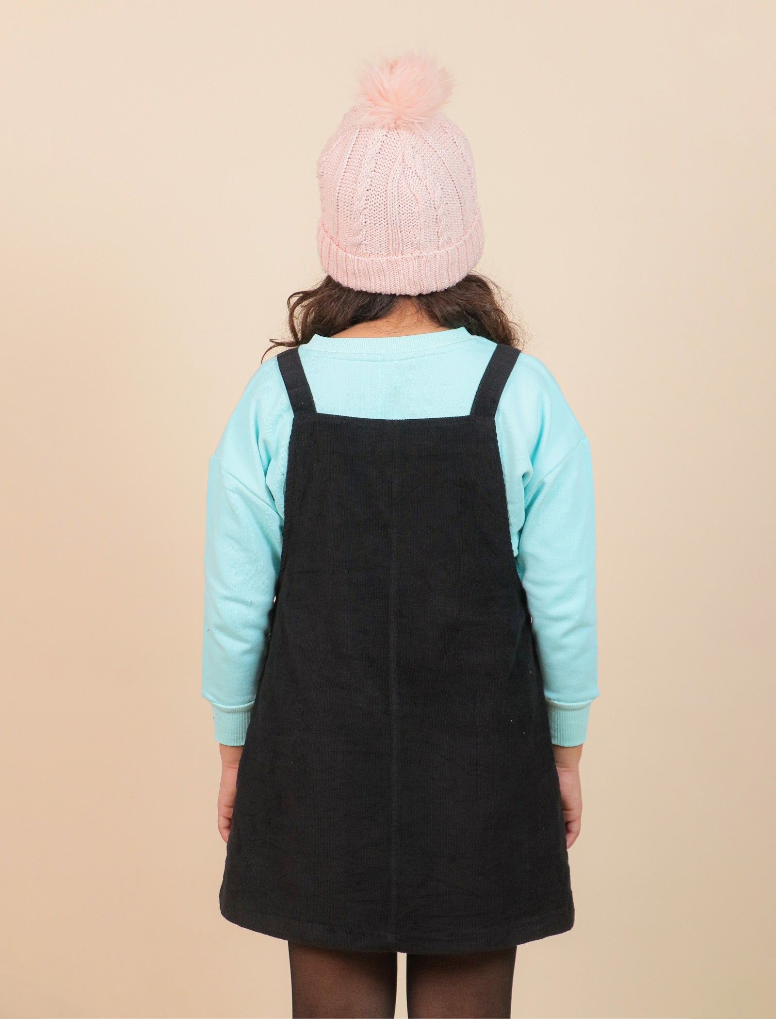 Girls wearing a black pinafore dress over a light blue long-sleeve shirt with a pink knit hat on a beige background