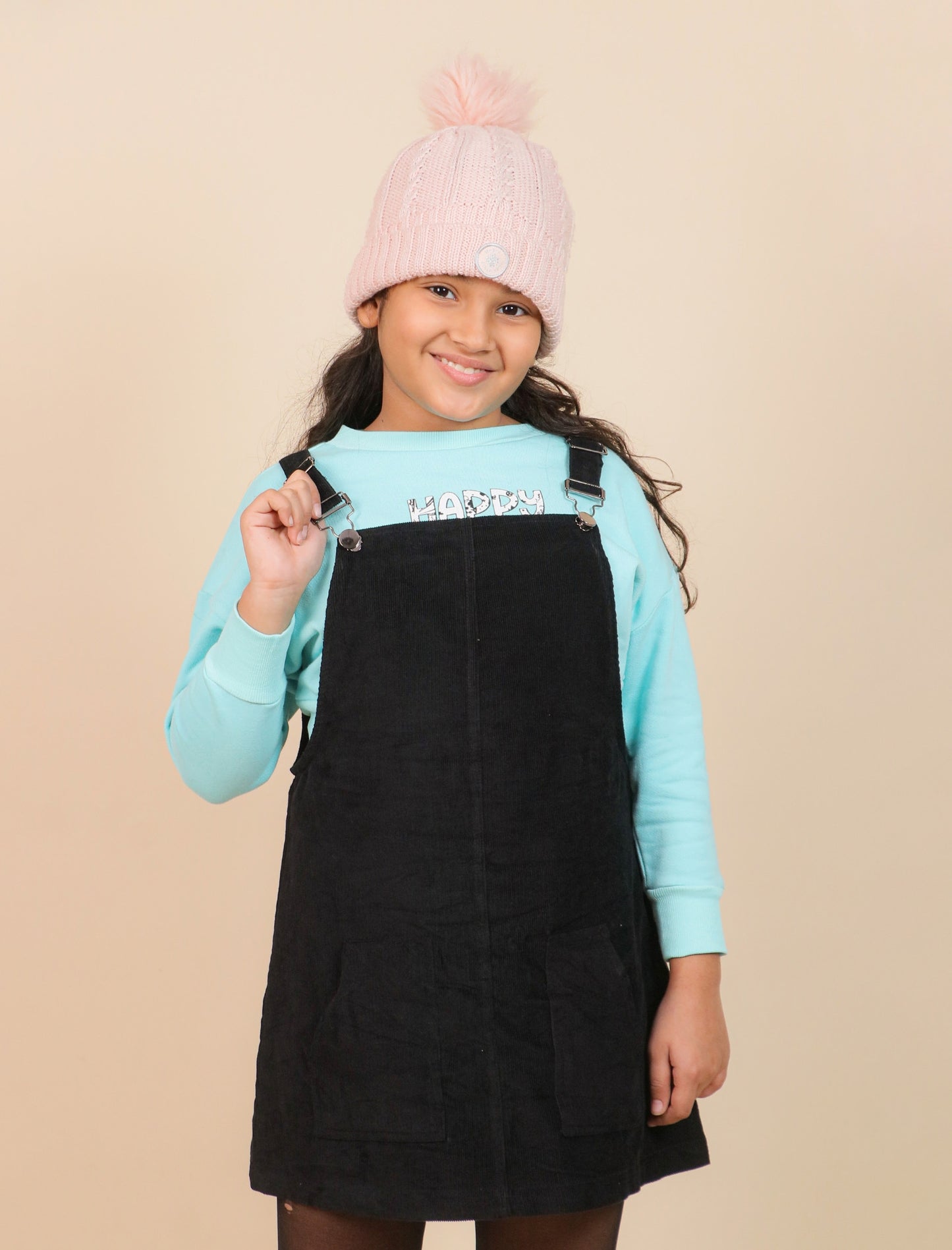 girls wearing a black pinafore dress over a light blue sweater with a pink beanie on a beige background