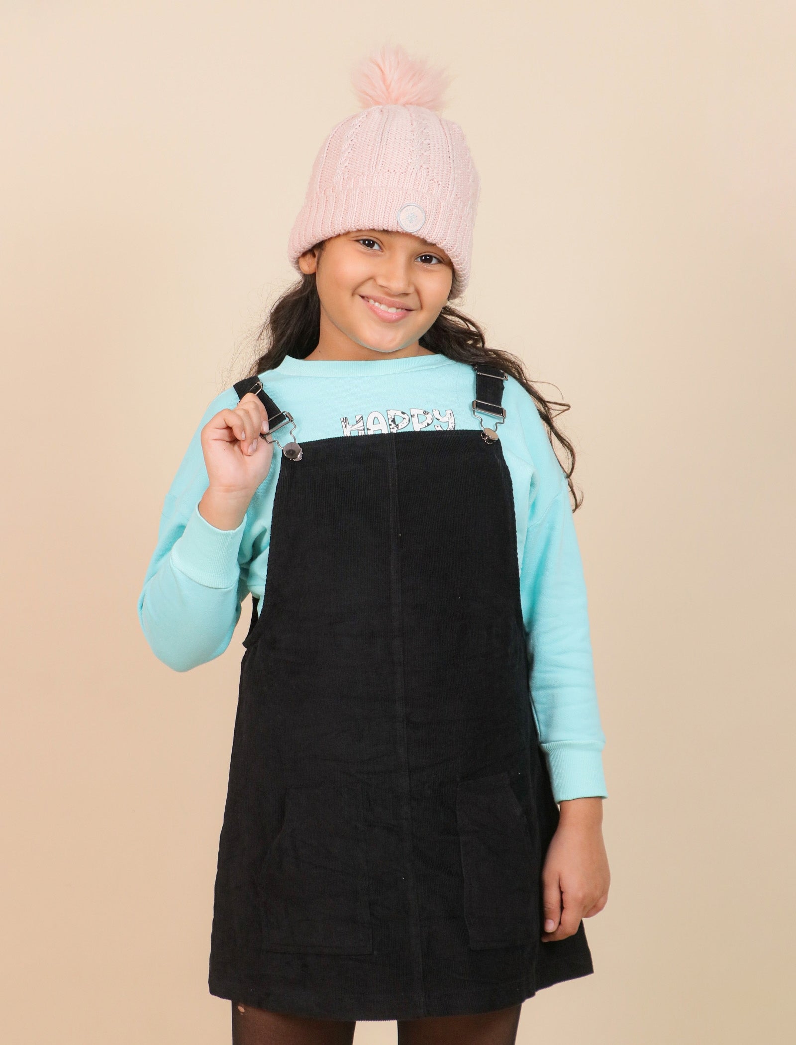 girls wearing a black pinafore dress over a light blue sweater with a pink beanie on a beige background